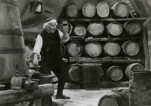 Scena del film "Don Buonaparte" - Regia Flavio Calzavara, 1941 - Figura intera di Ermete Zacconi che tiene un bicchiere di vino all'altezza degli occhi e lo osserva. Sullo sfondo, botti di vino.