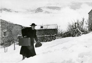 Scena del film "Don Camillo" - Regia Julien Duvivier, 1952 - Totale. Fernandel cammina sulla neve con una valigia nella mano destra e un fagottino nella sinistra.