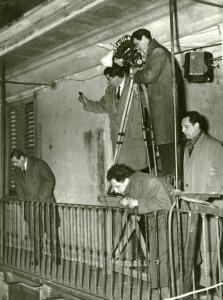 Sul set del film "La donna del giorno" - Regia Francesco Maselli, 1957 - Francesco Maselli si sporge, per guardare in basso, da un parapetto. Dietro di lui, un operatore con cinepresa in piedi su un cavalletto e altri membri della troupe.