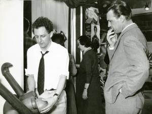 Sul set del film "La donna del giorno" - Regia Francesco Maselli, 1957 - Mezza figura di Francesco Maselli, a sinistra, con le maniche della camicia rimboccate, mentre sistema un oggetto di scena. Franco Fabrizi, a destra, di profilo, lo guarda.