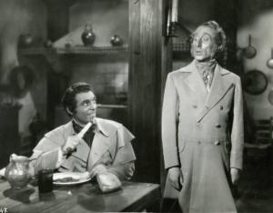 Scena del film "Il dottor Antonio" - Regia Enrico Guazzoni, 1937 - Mino Doro, seduto a tavola, porta alla bocca un coltello con infilzato del cibo e sorride a un attore non identificato in piedi accanto a lui.