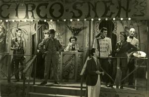 Scena del cortometraggio "Il domatore Suek" - Regia Piero Pierotti - Totale di un palco del circo. Sopra di esso, in piedi, un gruppo di donne e uomini con indosso costumi diversi. Gli attori hanno lo sguardo rivolto verso un uomo ai piedi del palco.