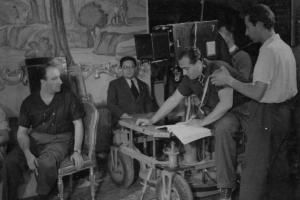 Sul set del film "Don Pasquale" - Regia Camillo Mastrocinque, 1940 - Giorgio Zambon, seduto sul carrello, è intento a leggere il copione. Intorno a lui il regista Camillo Mastrocinque e altri membri della troupe.