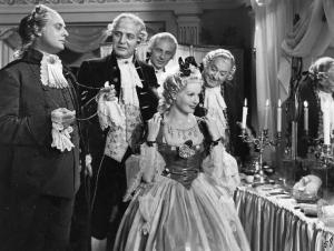 Scena del film "Don Pasquale" - Regia Camillo Mastrocinque, 1940 - Laura Solari, davanti a uno specchio, tiene una collana davanti al collo, come per provarla. Alle sue spalle, Gino Sabbatini. Attorno a loro, tre uomini guardano l'attrice.