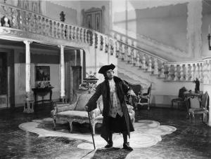 Scena del film "Don Pasquale" - Regia Camillo Mastrocinque, 1940 - Al centro di un atrio con scalinata, figura intera di Armando Falconi, che si appoggia a un bastone.