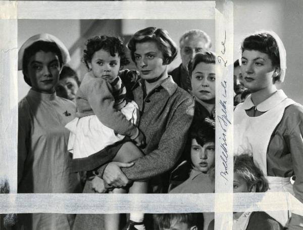 Scena del film "Europa 51" - Regia Roberto Rossellini, 1952 - Al centro, Ingrid Bergman con un'infante attrice non identificata in braccio, insieme a Giulietta Masina, a destra, rivolge lo sguardo a sinistra. Dietro, attori non identificati.