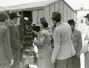 Sul set del film "Le due orfanelle" - Regia Carmine Gallone, 1942 - Il regista Carmine Gallone, a destra di spalle, con sigaretta in mano, osserva un operatore davanti alla macchina da presa. Attorno a loro, operatori non identificati.