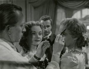 Sul set del film "Le due orfanelle" - Regia Carmine Gallone, 1942 - A sinistra, un operatore non identificato ritocca l'acconciatura di Alida Valli. Al centro, María Denis guarda la scena. Alle loro spalle, un attore non identificato osserva.