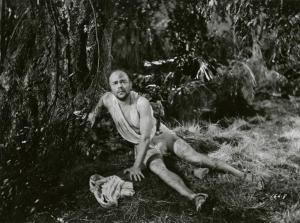 Scena del film "Le due tigri" - Regia Giorgio Candido Simonelli, 1941- Amedeo Trilli seduto, si appoggia con la mano sinistra al terreno e con la mano destra al tronco di un albero e guarda dritto davanti a sé.