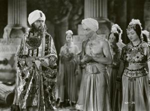 Scena del film "Le due tigri" - Regia Giorgio Candido Simonelli, 1941- Cesare Fantoni, sulla sinistra, osserva un attore sorridente non identificato al centro. Sulla destra e alle loro spalle, sono presenti attrici non identificate.