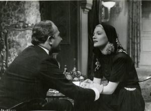 Scena del film "È caduta una donna" - Regia Alfredo Guarini, 1941 - Luigi Pavese, a sinistra di spalle, stringe le mani di Isa Miranda, a destra, che lo osserva intensamente. Entrambi sono seduti a un tavolino.