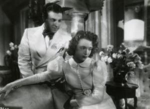 Scena del film "Ecco la felicità!" - Regia Marcel L'Herbier, 1940 - In primo piano, Micheline Presle seduta su un divano, rivolge lo sguardo verso destra, mentre Ramon Novarro, in piedi alle sue spalle, la osserva.