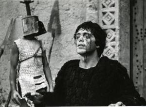 Scena del film "Edipo Re" - Regia Pier Paolo Pasolini, 1967 - Franco Citti ha gli occhi chiusi e truccati e le braccia semi-alzate. Intanto, dietro di lui, un attore non identificato con un elmo sul capo, rivolge lo sguardo verso destra.