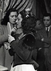 Sul set del film "Elisir d'amore" - Regia Amleto Palermi, 1941 - Margherita Carosio, in primo piano, si guarda in uno specchio, mentre un'operatrice non identificata la pettina. In secondo piano, altri operatori non identificati.