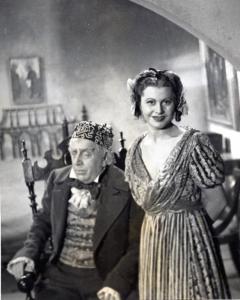 Sul set del film "Elisir d'amore" - Regia Amleto Palermi, 1941 - A sinistra, Armando Falconi, seduto su una sedia, guarda verso sinistra, mentre Gemma D'Alba, a destra in piedi, osserva dritto davanti a sé.