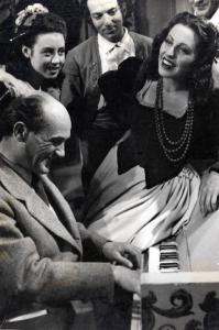 Sul set del film "Elisir d'amore" - Regia Amleto Palermi, 1941 - In primo piano il regista Amleto Palermi suona un pianoforte su cui è seduta Margherita Carosio. In secondo piano attori e attrici non identificati osservano.
