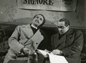 Sul set del film "La fabbrica dell'imprevisto" - Regia Jacopo Comin, 1942 - Maurizio D'Ancora seduto guarda verso l'alto con le mani conserte mentre, a destra, il regista Jacopo Comin legge un plico di fogli.