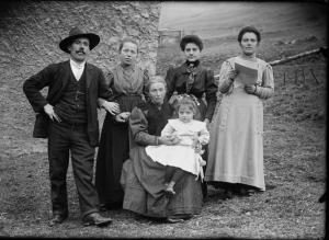 Val Brembana. Ritratto di famiglia con casa rurale e campo sullo sfondo. Donna con giornale in mano