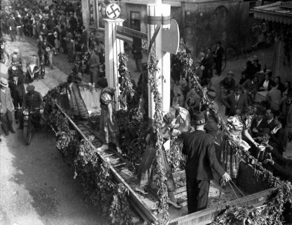 Broni - Piazza Garibaldi - VIII Festa dell'uva - Carro folcloristico con il Fascio littorio e lo stemma della Germania nazista (Terzo Reich) - Giovani donne in costume distribuiscono tazze di vino - Folla