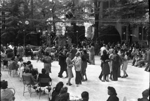 Salice Terme - IX Festa nazionale dell'uva - Coppie di uomini e donne danzano - Gruppo di persone sedute ai tavolini - Sullo sfondo in posizione sopraelevata l'orchestra con uomini che suonano strumenti musicali
