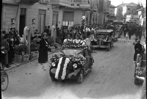 Salice Terme - IX Festa nazionale dell'uva - Sfilata di un'autovettura addobbata con fiori e in piedi tre giovani donne in abiti tradizionali, seguita da altre vetture - Una folla è disposta lungo la strada