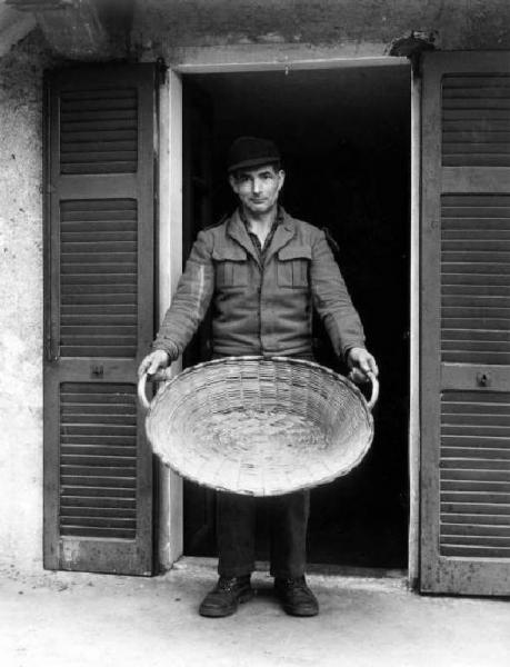 Il cesto per le olive. Uomo in piedi contro una porta-finestra aperta regge grande cesto in vimini ("la val") per scuotere le olive per la prima monda. Luigi Di Battista.