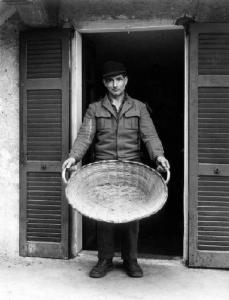 Il cesto per le olive. Uomo in piedi contro una porta-finestra aperta regge grande cesto in vimini ("la val") per scuotere le olive per la prima monda. Luigi Di Battista.