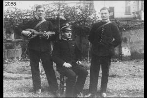 Ritratto di Renzo Vitari, da Fuipiano Valle Imagna. Tre uomini in divisa in posa in un cortile. Uno di loro suona il mandolino.