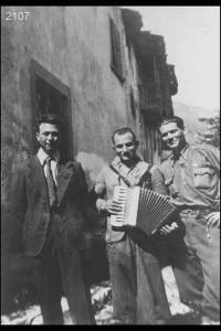 Egidio Angiolini, Ambrogio Manzoni detto 'scinca',   Antonio Vitari detto 'marsenta'. Posa davanti a edifici di campagna. L'uomo al centro suona la fisarmonica.