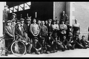 Operai in posa davanti all' ingresso di capannone di fabbrica. A terra appoggiato alla bicicletta Giovanni Rota.
