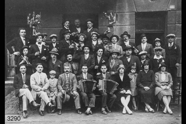 Emigranti di Valsecca e S.Omobono - Posa in esterno. - Uomini e donne con fisarmoniche e bottiglie di vino.