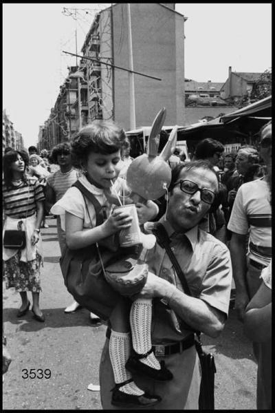 Festa del Giglio al Quartiere Lorenteggio. Gente del Pubblico. Uomo con bambina in braccio. La bambina ha un pupazzo gonfiabile a forma di coniglio e sta bevendo una bibita in lattina con la cannuccia.