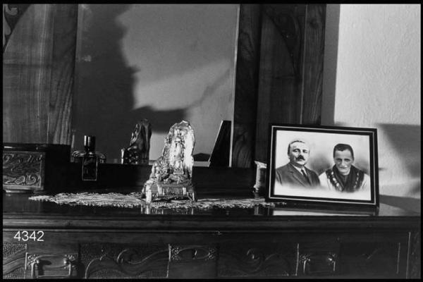 Interno di abitazione. Fotografia di famiglia, Madonna di Lourdes - soprammobile, centrino e bottiglia di profumo sul piano di un comò