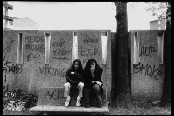 Coppia di ragazze su di una panchina. Sullo sfondo muro con graffiti.