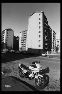 Quarto Oggiaro.  Una via del quartiere: in primo piano una moto, sullo sfondo alcuni  condominii.