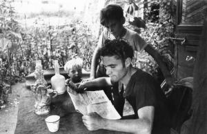 Milano. Baracche. Un uomo ad un tavolo legge un giornale. A fianco, due ragazzini.