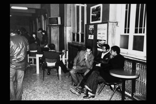 Eritrei a Milano. Bar. Teatro Officina. Persone in piedi e ai tavoli. Sulla destra tre eritrei discutono seduti.