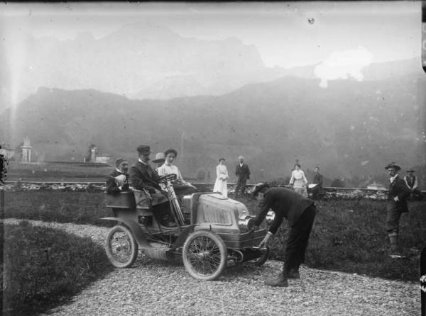 Automobile Rovetta in giardino. Un uomo avvia a manovella una automobile su cui siede una giovane coppia e, dietro, un vecchio che regge un imbuto.