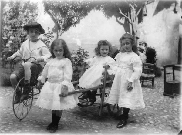 Tre bambine in abiti eleganti e un bambino vestito alla marinara giocano in un giardino con un carrettino e un triciclo.