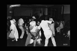 Eritrei a Milano. Gruppo che danza al centro del locale. Tutt'intorno eritrei seduti accompagnano le danze battendo le mani.