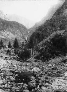 Al fonte del Gheno (prima della diga) - Due uomini e una donna in tenuta escursionistica attraversano un piccolo torrente di montagna. - Inquadratura panoramica, in verticale, della valle con cascatella in quota.