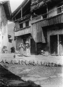 Piazza di Pianezza - Slargo con edifici rustici;  al centro dell'inquadratura giovane signora con ombrellino accanto a contadinella con gerla sulle spalle.