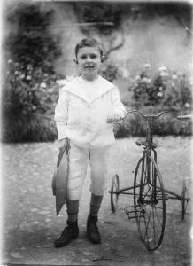 Carlo col triciclo a Brescia. Bambino in posa frontale, a figura intera, in giardino. Accanto a lui un triciclo in ferro.