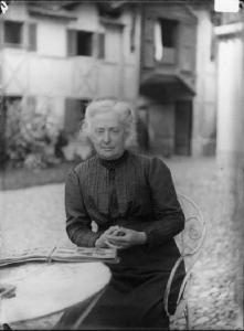 Signora Virginia Danieli Furlotti. Donna anziana seduta ad un tavolino di un cortile con giardino. Sul tavolino, un album fotografico aperto.
