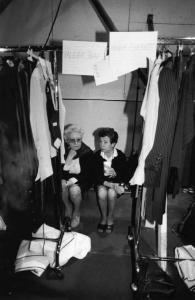 Coppia di assistenti addette a una sfilata di moda sedute nei camerini delle modelle tra due appendiabiti, in un momento di pausa.