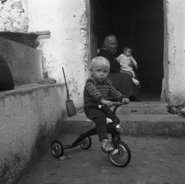Melissa (Crotone) - Bambino su triciclo e anziana donna con bambino in braccio sull'uscio di una casa