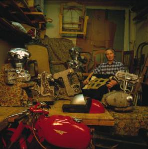 Uomo adulto in posa tra pezzi meccanici di motociclette - Scorcio di moto d'epoca in primo piano