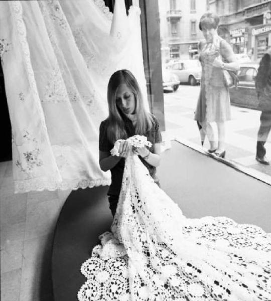 Giovane donna con tovaglia ricamata in una vetrina di negozio - una signora guarda dall'esterno