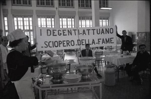 Sciopero della fame dei ricoverati presso il Sanatorio di Vialba: interno della mensa del centro ospedaliero. In primo piano una cuoca che riempie un piatto nella sala da pranzo vuota. Dietro di lei, uno striscione comunica lo sciopero della fame