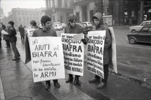 Manifestazione per il Biafra: tre ragazzi leggono un volantino e portano al collo cartelli di protesta contro le armi e il genocidio in Nigeria